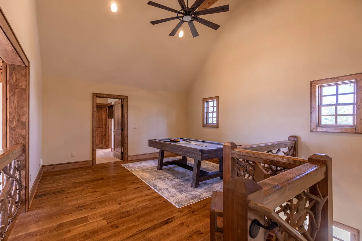 A spacious loft area with a pool table as its centerpiece, flanked by wooden railings and bathed in natural light streaming through windows. The setting conveys an inviting atmosphere ideal for social gatherings, with a balance of playfulness and relaxation.