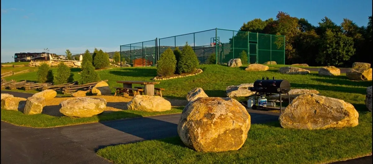 The scene shows a relaxed, friendly outdoor gathering space with large boulders, a picnic area, tennis court, and grill. People are seen enjoying barbecues, playing tennis, and socializing in a natural setting.