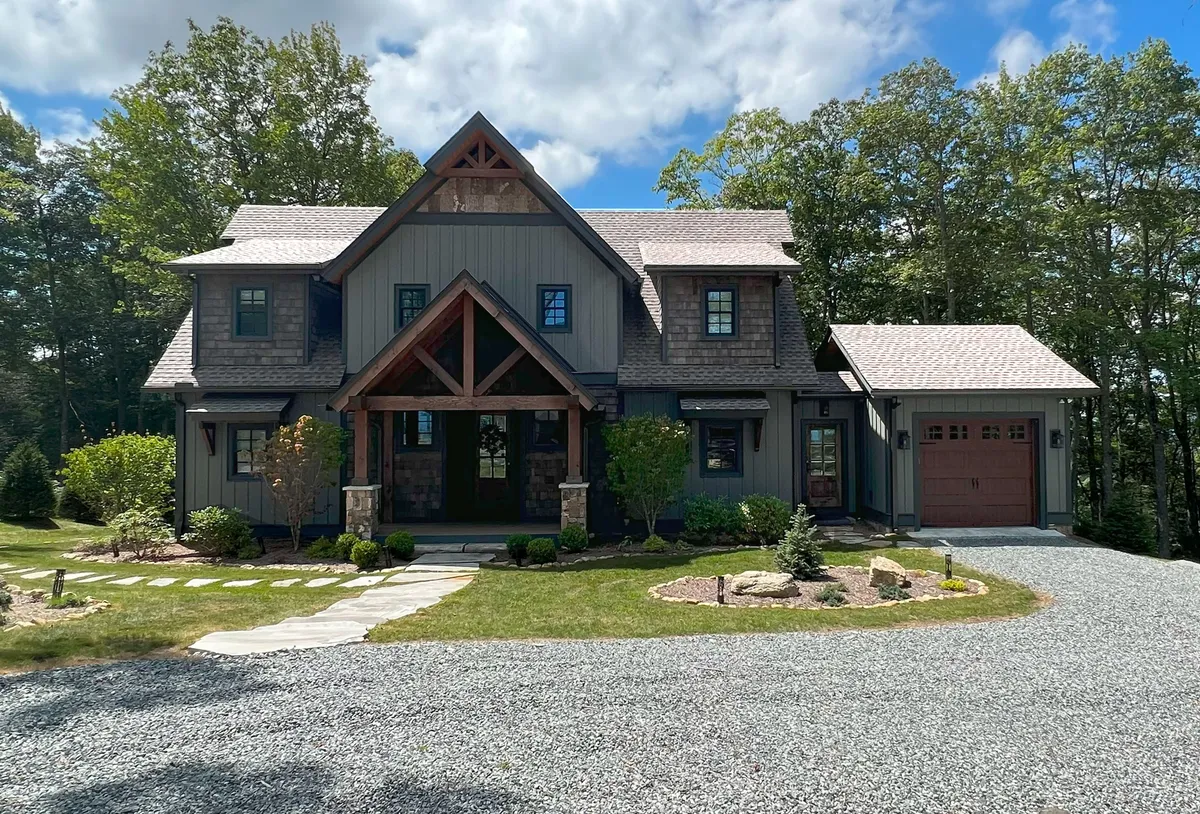 The image showcases the exterior of Antler Ridge, with a spacious front porch framed by rustic wooden beams and elegant stonework, surrounded by lush landscaping. The setting is tranquil, offering plenty of space for outdoor relaxation and gatherings, and hints at breathtaking mountain views.