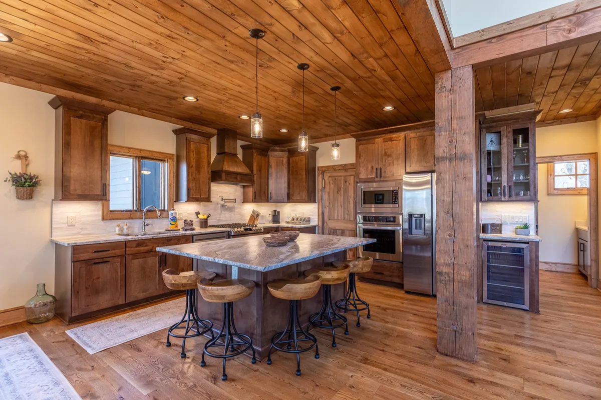 A contemporary kitchen with a large island, wooden cabinetry, and stainless steel appliances, bathed in natural light from nearby windows. Pendant lights add to the inviting atmosphere, and the space invites both meal preparation and casual gathering.