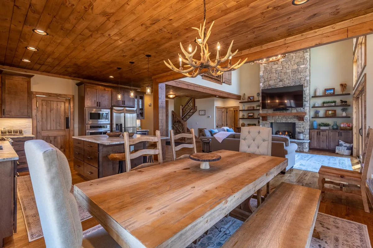 A cozy dining area flows seamlessly into the great room, featuring a dining table beneath an antler chandelier, a view of the fireplace, and wooden cabinetry. The space is set for shared meals and special occasions, embodying warmth and togetherness.