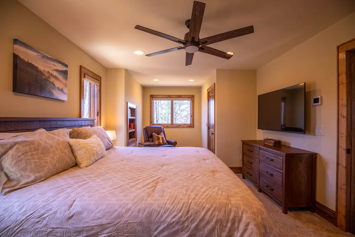 A spacious king suite featuring a large bed adorned with crisp white linens, complemented by a ceiling fan, windows letting in natural light, decorative artwork, and a wall-mounted television. The room exudes a tranquil and comfortable atmosphere, ideal for relaxation and restful nights.