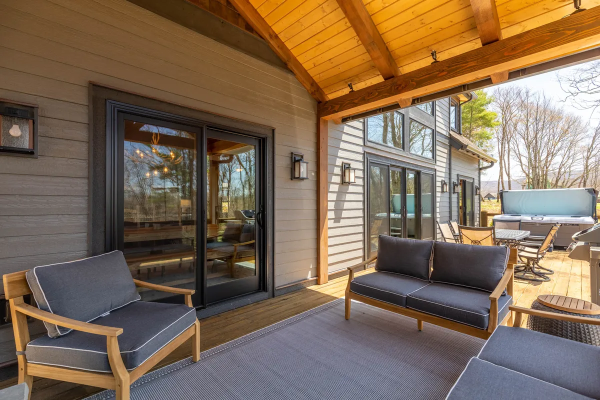 A spacious outdoor seating area is shown, with comfortable chairs and a large dining table arranged on a deck. The hot tub is tucked in the corner, surrounded by lush trees and set against the backdrop of large windows that look into the home. The setting is ideal for group gatherings, outdoor meals, and relaxing moments in nature.