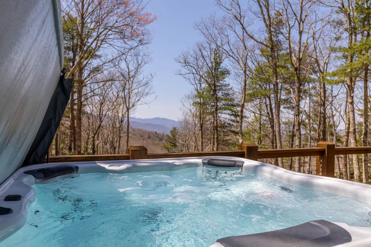 A tranquil exterior shot showing a bubbling hot tub nestled on a deck, framed by stunning mountain views and surrounding trees. The setting sun casts a warm glow over the landscape, evoking a peaceful end to a day of adventure.