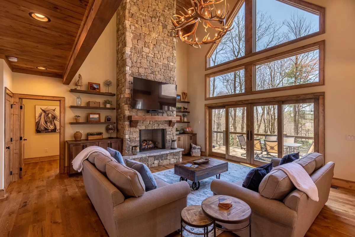 A spacious great room with a majestic stone fireplace as the focal point. Large, comfortable sofas are arranged for conversation or relaxation. Expansive windows let in abundant natural light, while a chandelier hangs from the soaring vaulted ceilings, creating a sense of openness and warmth.