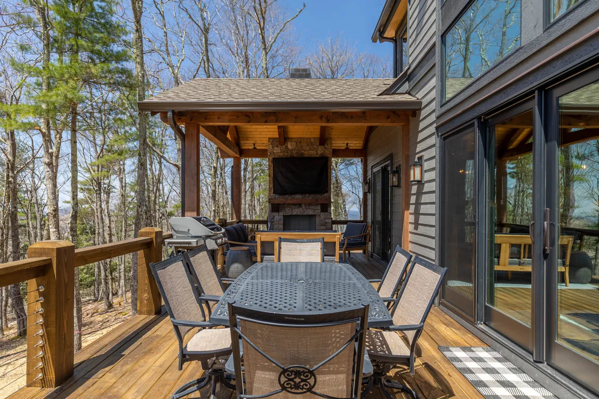 A deck featuring a large outdoor dining table and an impressive stone fireplace, surrounded by trees, is shown. The space is set up for outdoor dining, barbecues, and cozy fire-lit evenings, with the natural setting creating a tranquil backdrop.