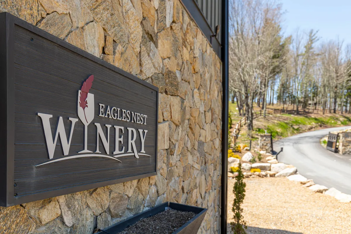 A welcoming stone path leads to the Eagles Nest Winery sign, set against a rustic stone wall and surrounded by greenery. The scene is inviting and distinctly tied to the local wine culture, with a gentle pathway hinting at leisurely strolls and the promise of wine tasting experiences.