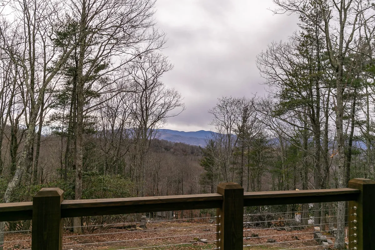 A sweeping view from the deck, with a wooden railing framing distant mountains and treetops. The tranquil mountain landscape provides a sense of retreat and offers opportunity for guests to relax, take in the scenery, or enjoy morning coffee outdoors.