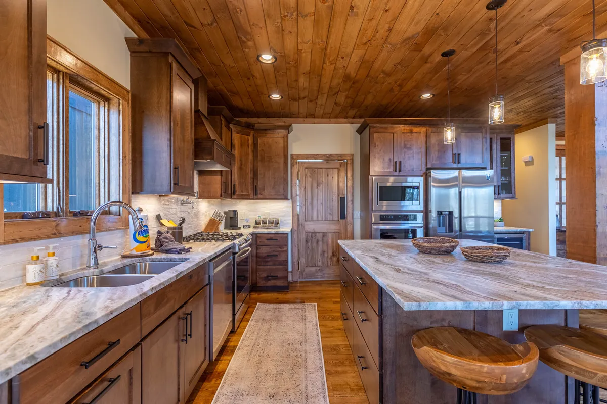 A sunlit kitchen with a prominent island, gleaming stainless steel appliances, wooden cabinetry, and natural light streaming in from the windows. The kitchen layout is spacious, with clear sightlines to the adjacent entertaining spaces.