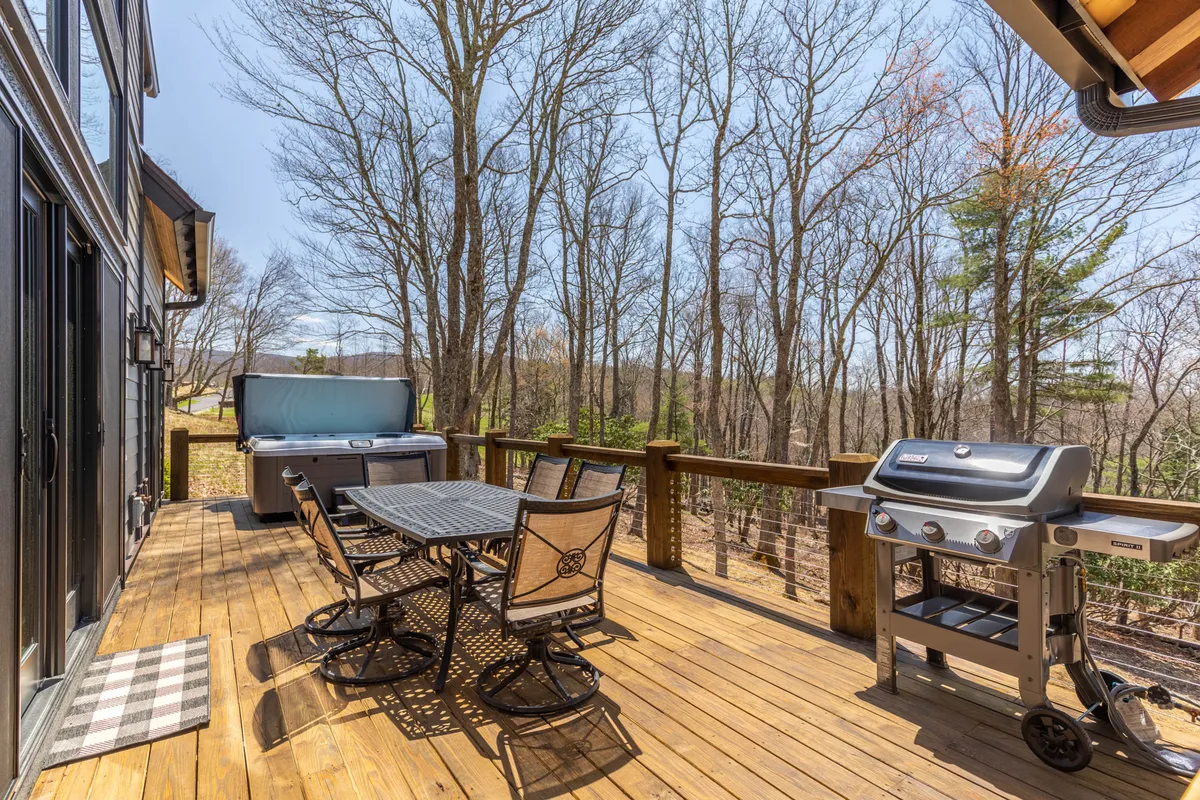 An expansive outdoor deck featuring a dining table with chairs, a grill, a hot tub, and surrounded by trees. The area is set up for socializing, outdoor meals, and relaxation, with the hot tub and seating suggesting both comfort and enjoyment of nature.